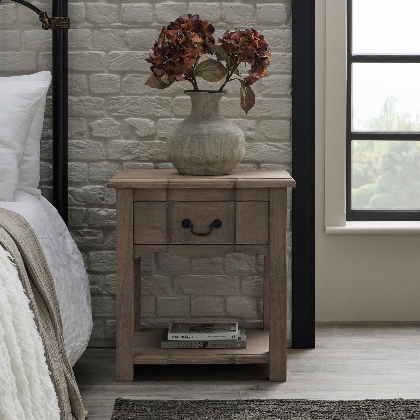Wooden nightstand with a vase of flowers next to a bed in a bedroom.