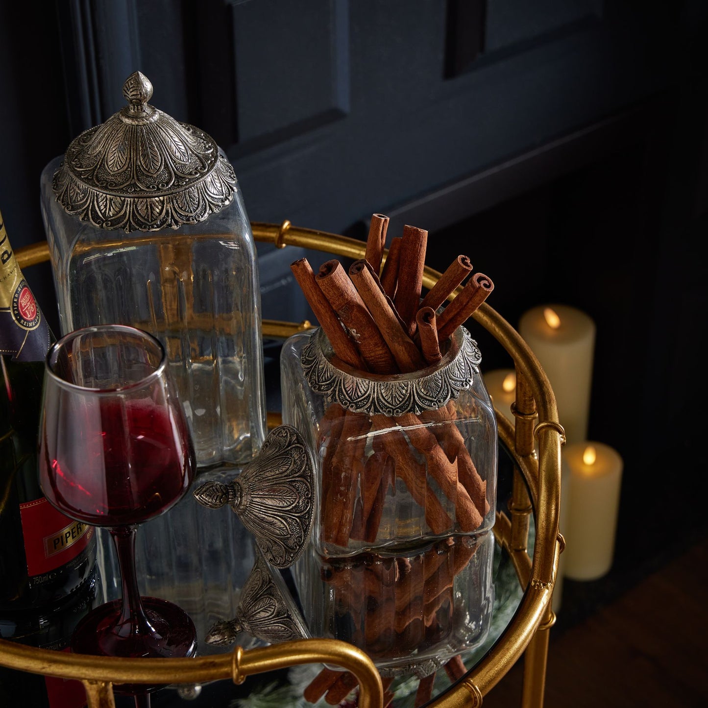 Decorative glass jar with cinnamon sticks on a gold tray with a glass of red wine and bottle.