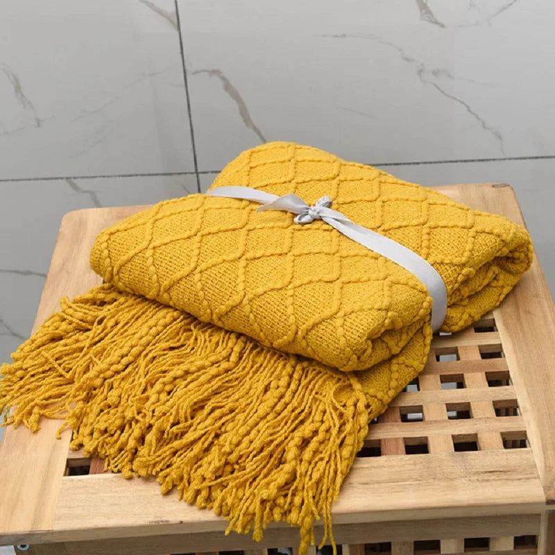 Yellow knitted blanket with tassels on a wooden stool against a gray tiled wall.