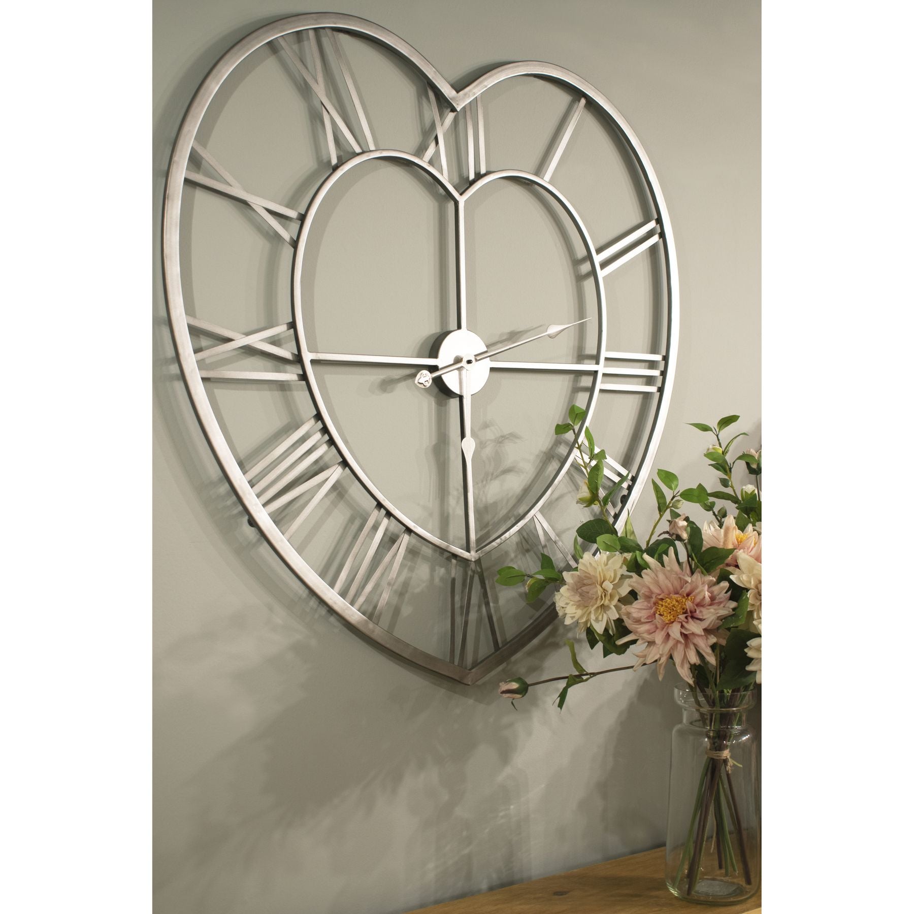 Heart-shaped wall clock with Roman numeral design on a light gray wall, accompanied by a vase of flowers.