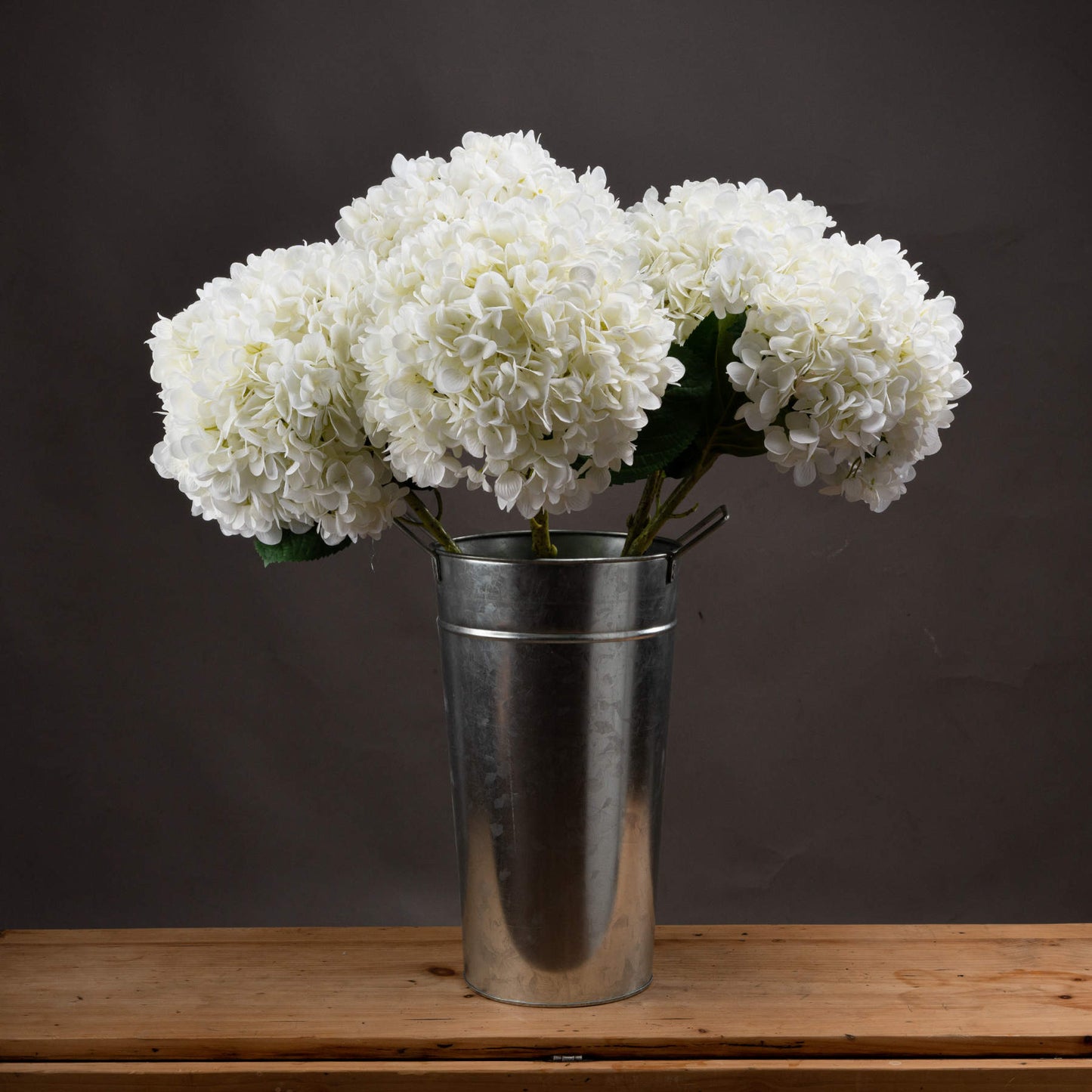 White hydrangeas in a silver vase on a wooden surface with a dark background