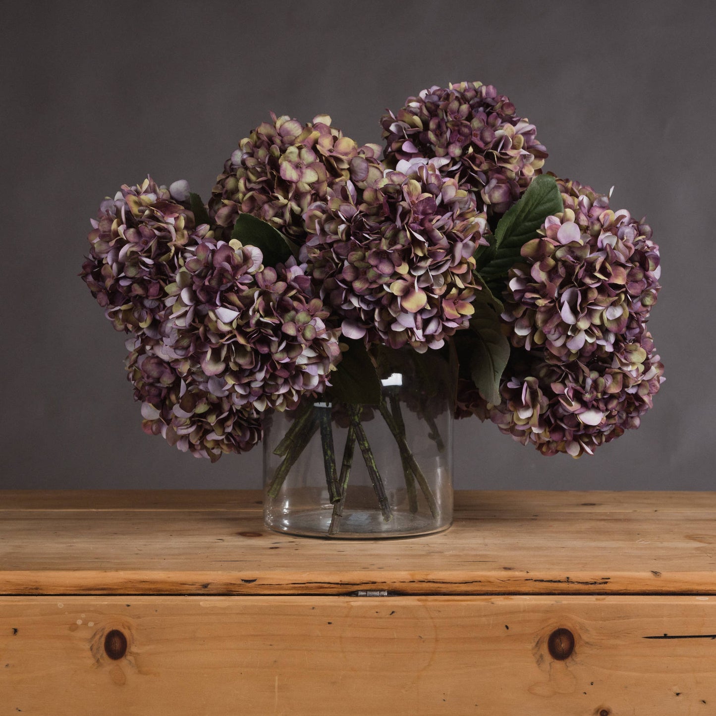 Bouquet of purple hydrangeas in a clear vase on a wooden surface with a gray background