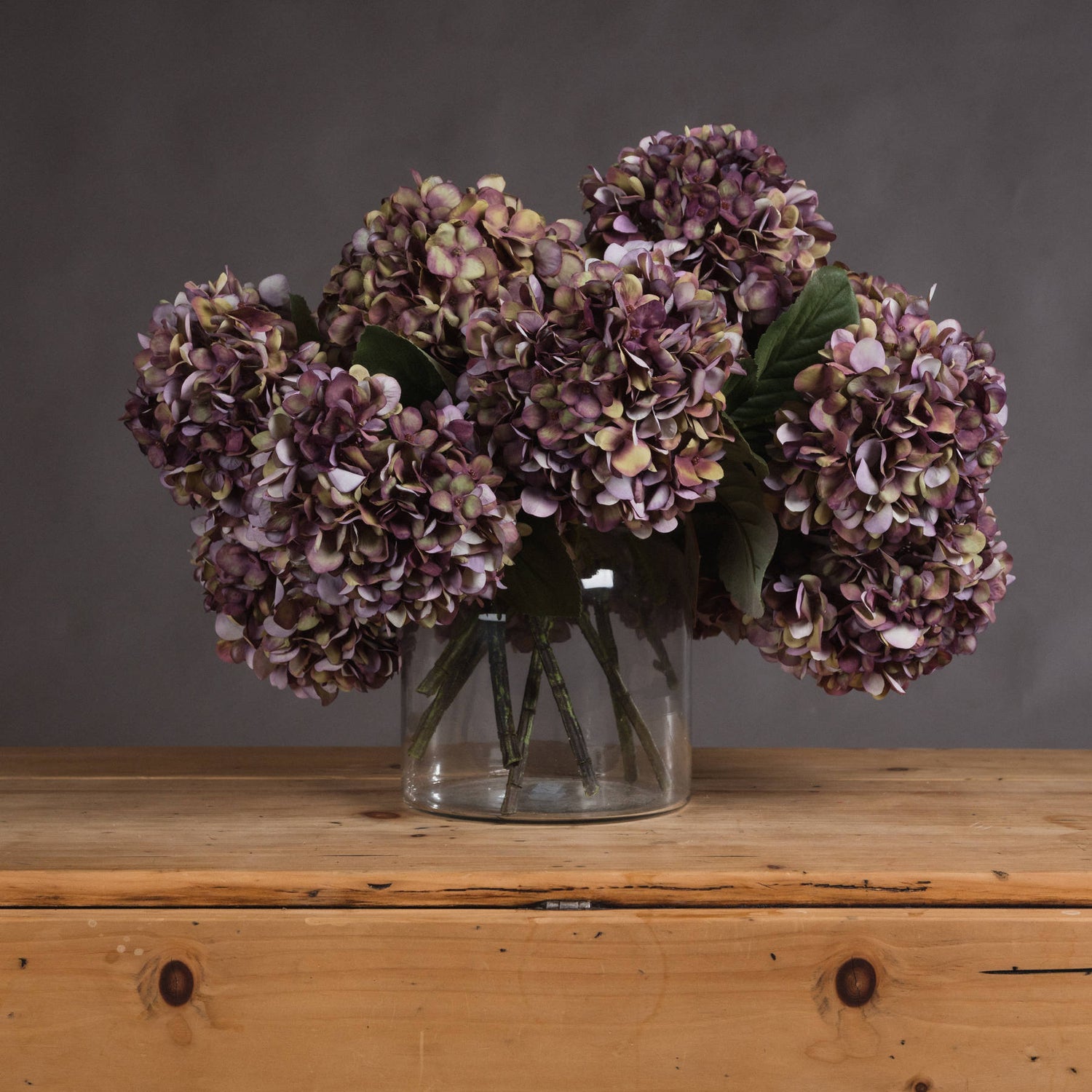 Bouquet of purple hydrangeas in a clear vase on a wooden surface with a gray background
