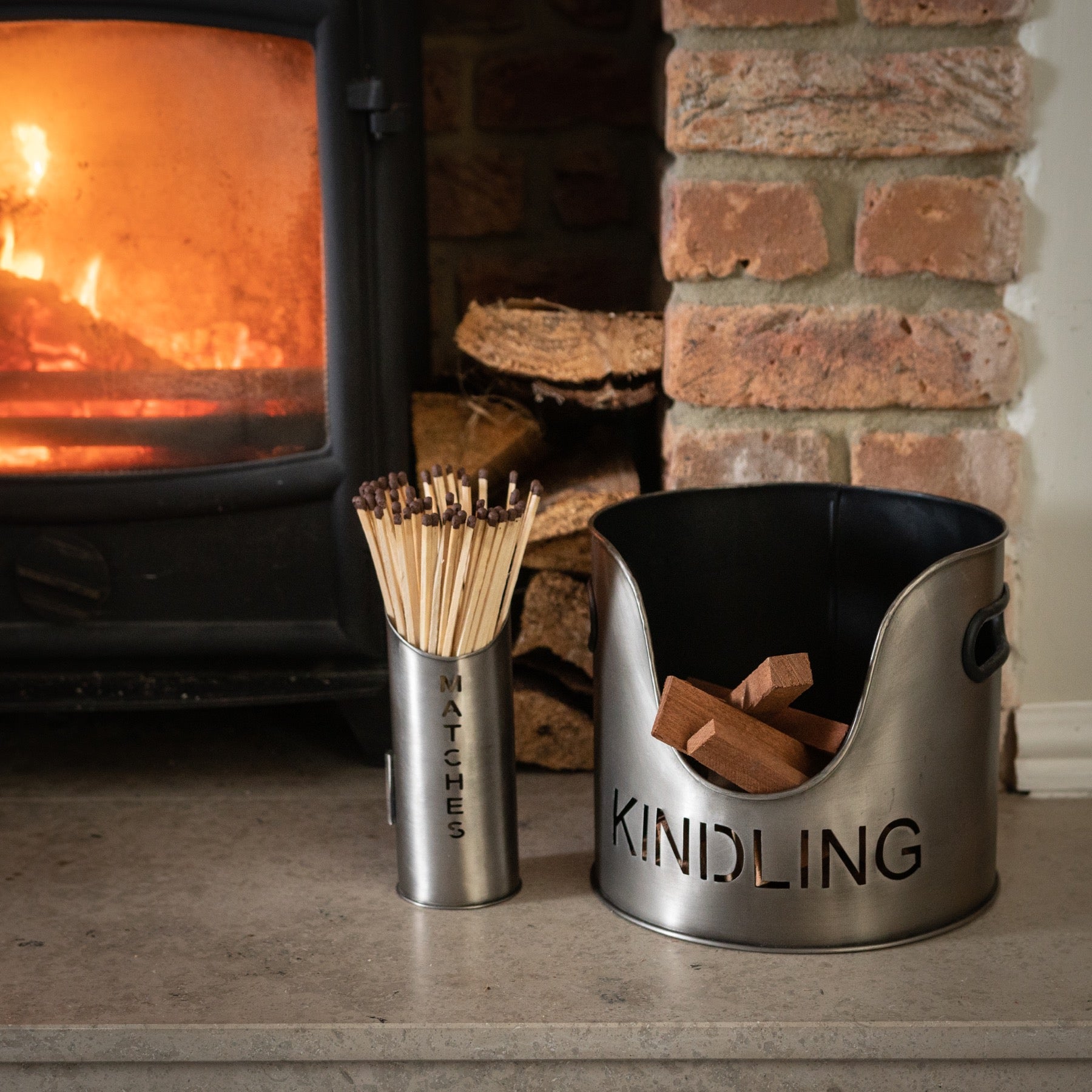 Two metal containers labeled 'Matches' and 'Kindling' in front of a lit fireplace.