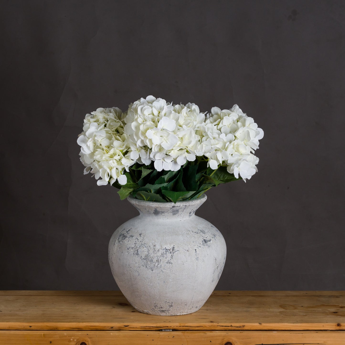 White hydrangeas in a textured vase on a wooden surface with a dark background