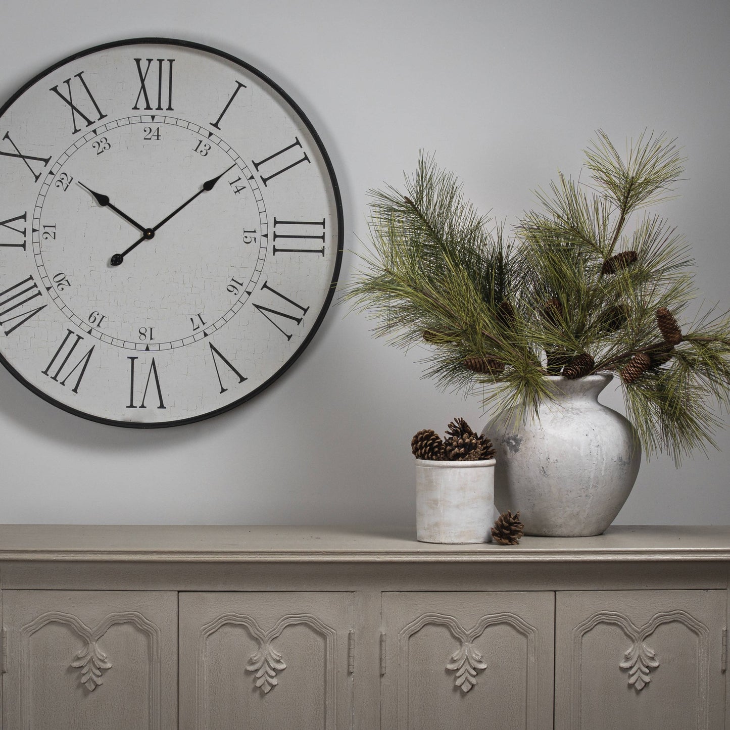 Decorative clock on a wall with a vase of greenery on a cabinet.