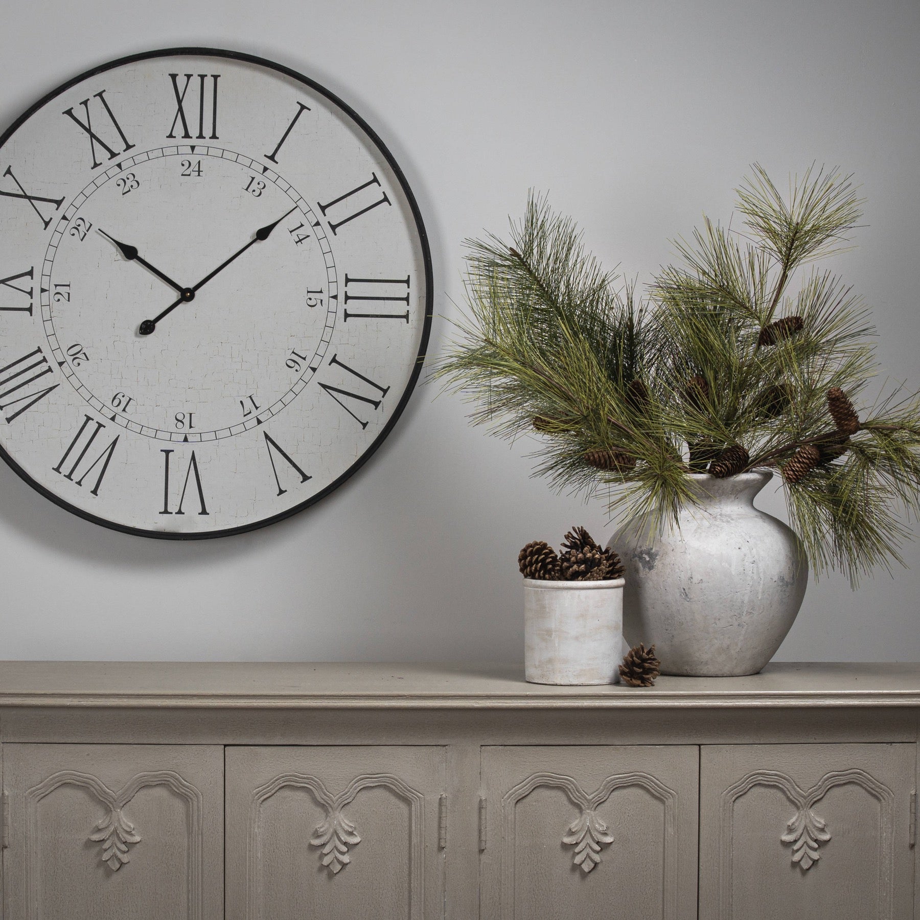 Decorative clock on a wall with a vase of greenery on a cabinet.