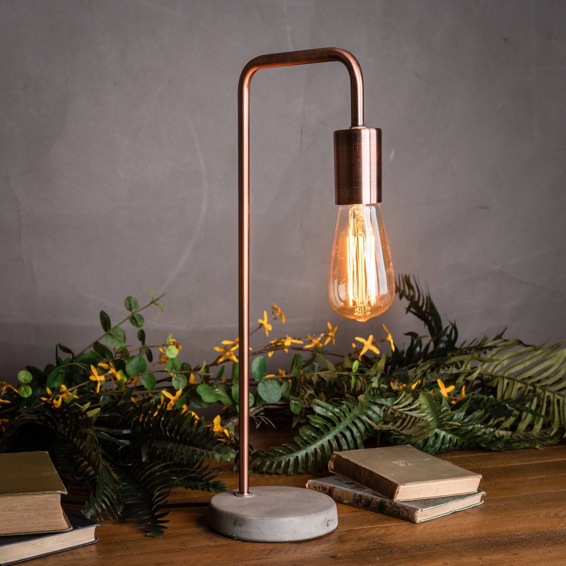 Copper table lamp with a lit bulb on a wooden surface with books and greenery.