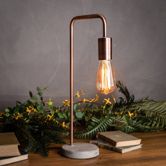 Copper table lamp with a lit bulb on a wooden surface with books and greenery.