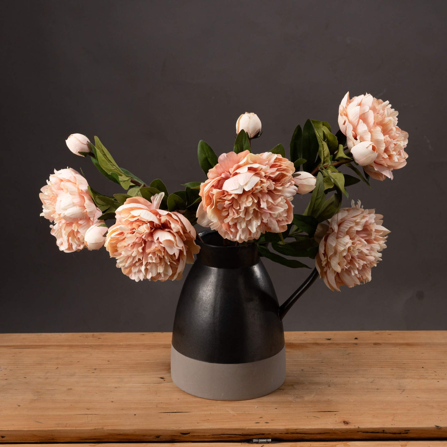 Black vase with peach-colored flowers on a wooden surface against a dark background