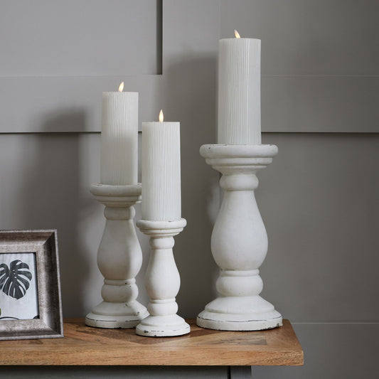Three white candle holders with candles on a wooden surface against a gray wall.
