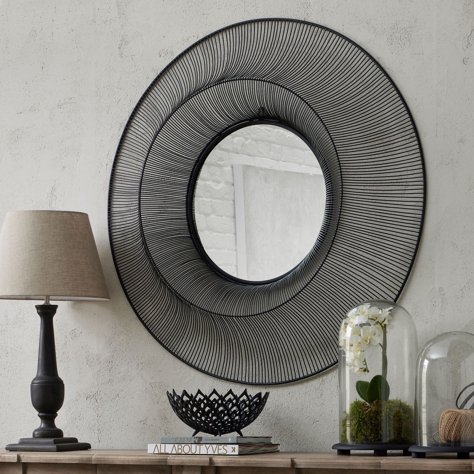 Decorative wall mirror with concentric circles on a neutral wall with a lamp and decorative items below.