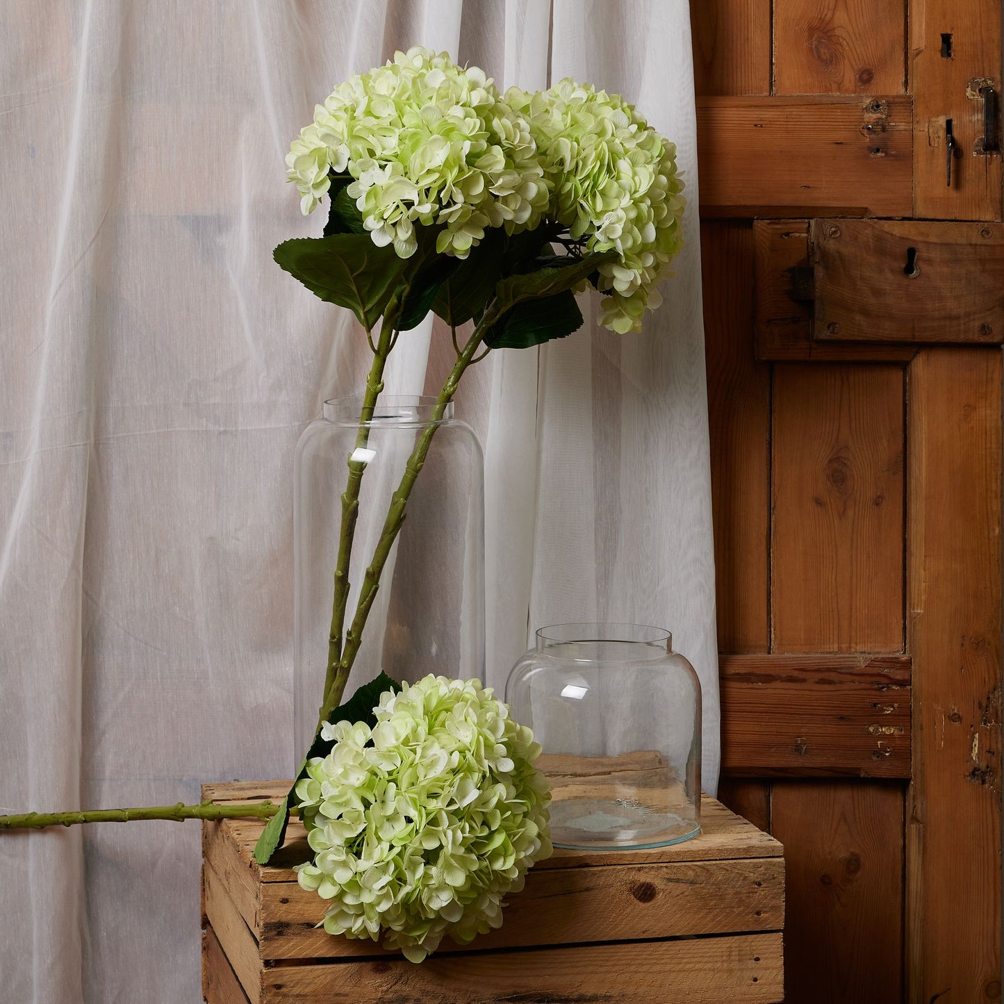 Green hydrangeas in a clear vase on a wooden crate with a wooden cabinet in the background.