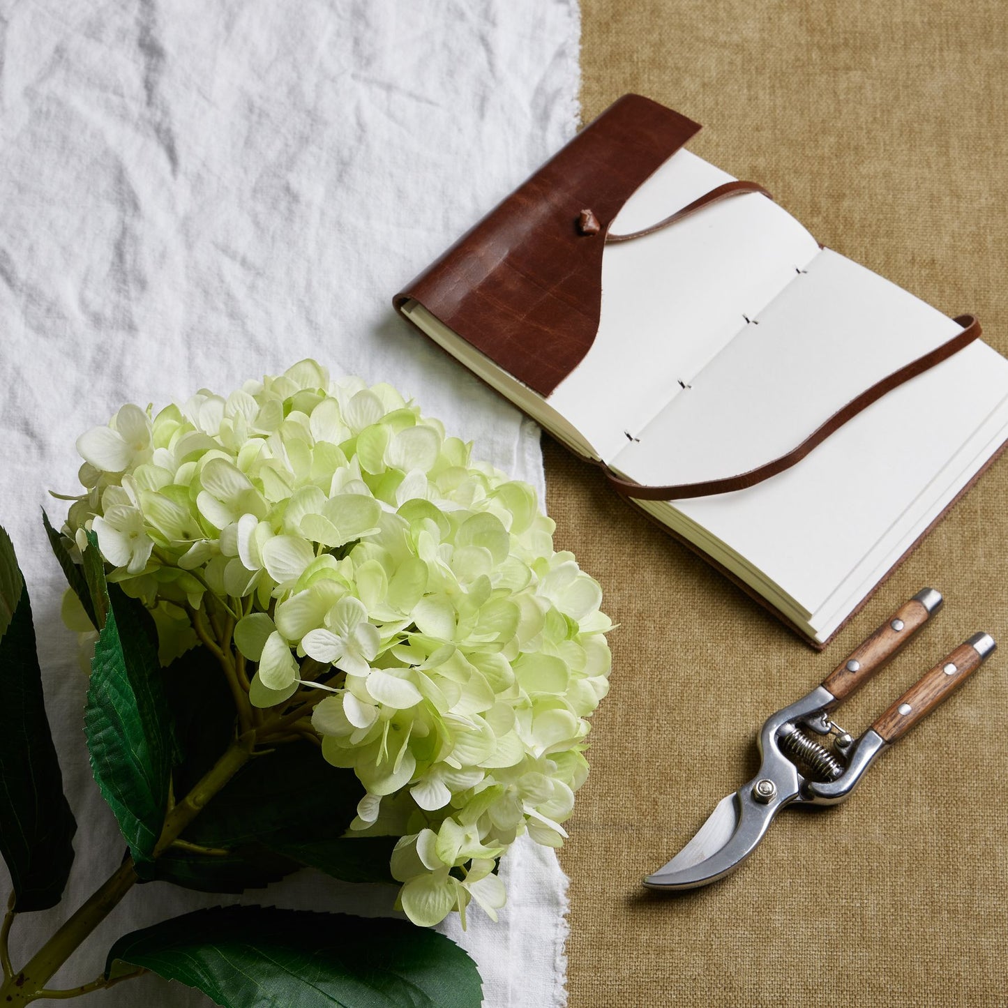 Open notebook with brown leather binding, green hydrangea flower, and garden shears on a textured surface.