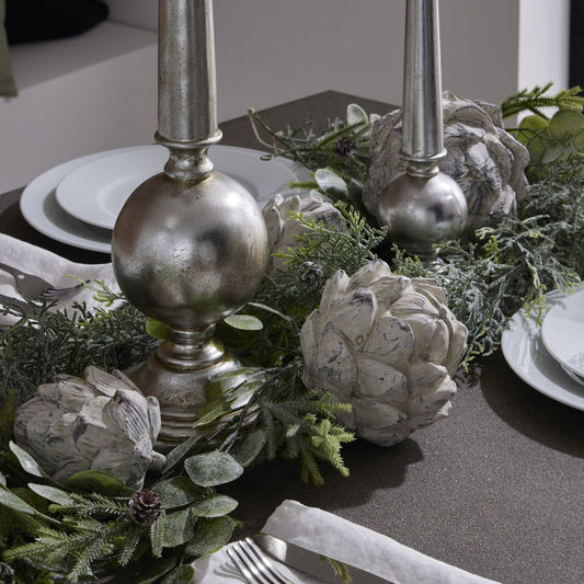 Decorative table setting with silver candlesticks, artichoke ornaments, and greenery.