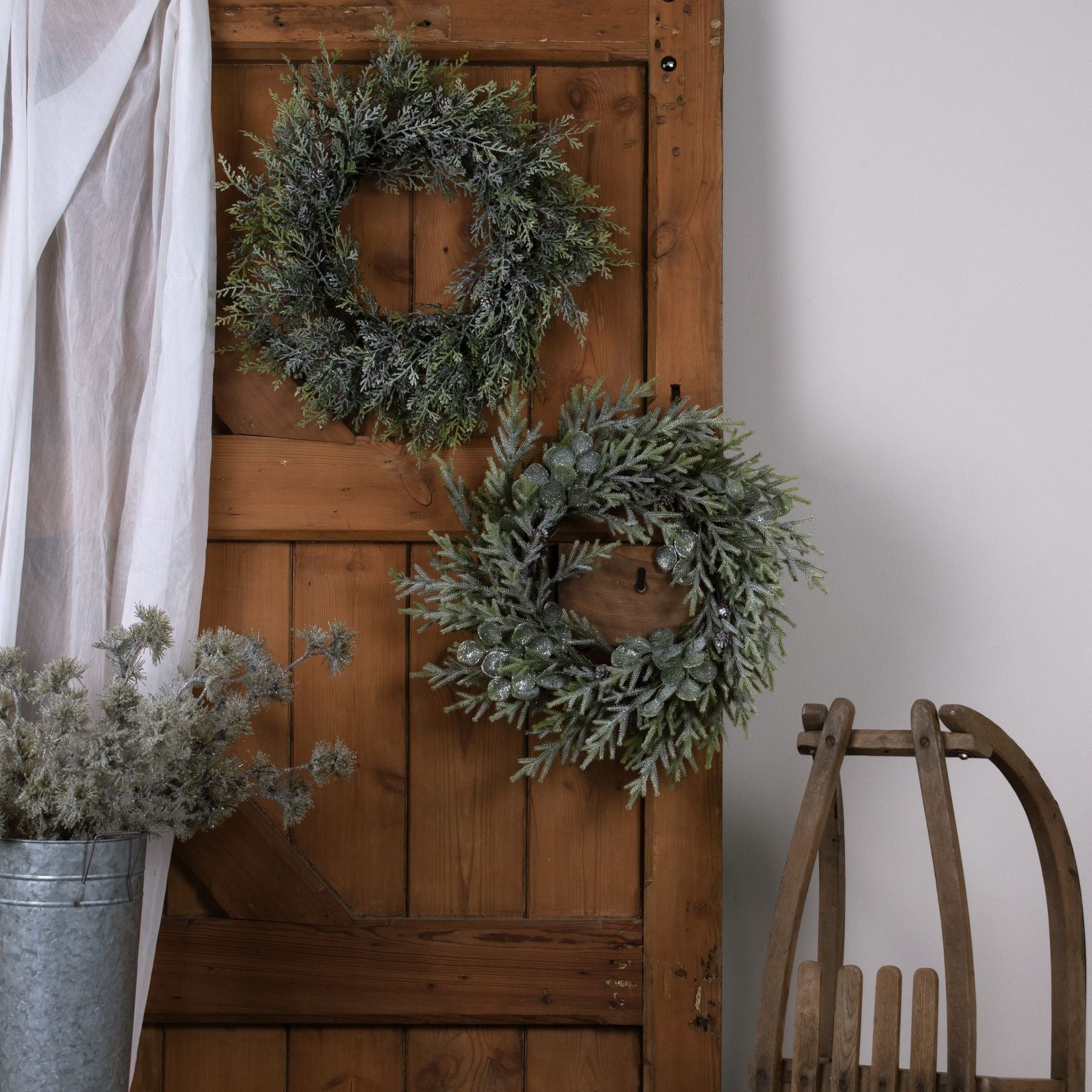 Two green wreaths on a wooden door with a wooden chair and plant in the foreground.