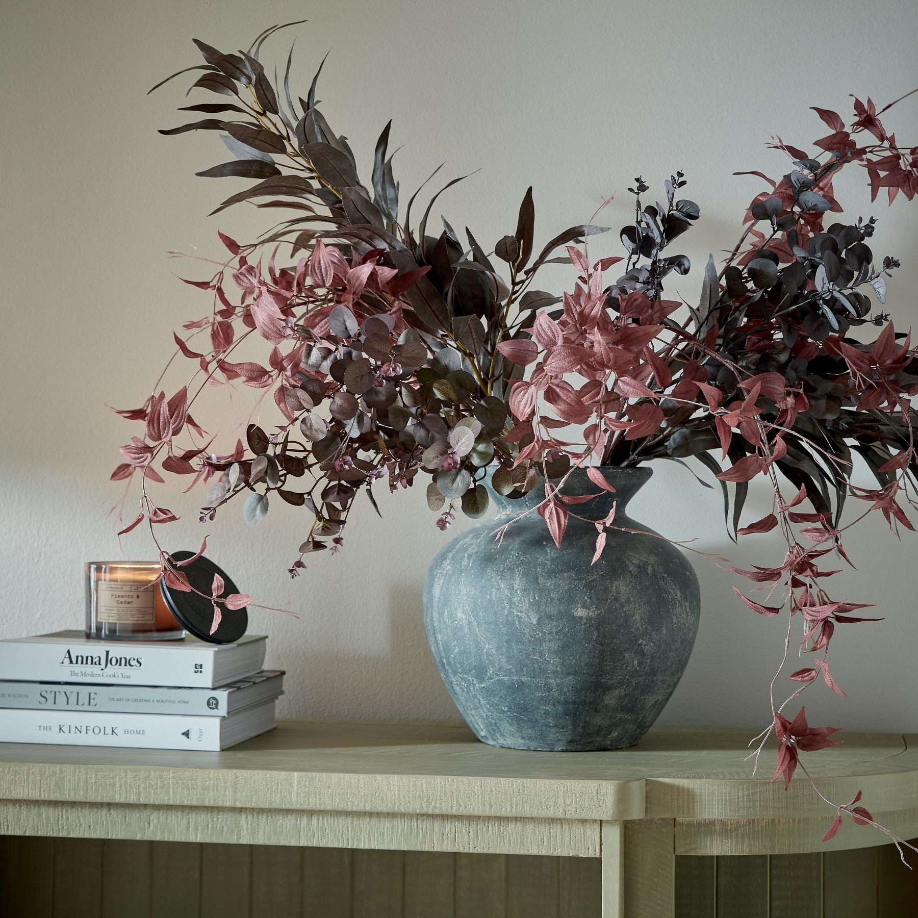 Decorative vase with pink and brown leaves on a wooden surface with books and a candle.