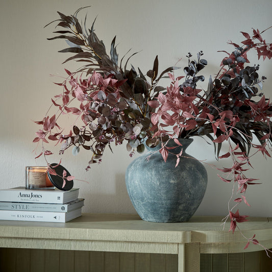Decorative vase with pink and brown leaves on a wooden surface with books and a candle.