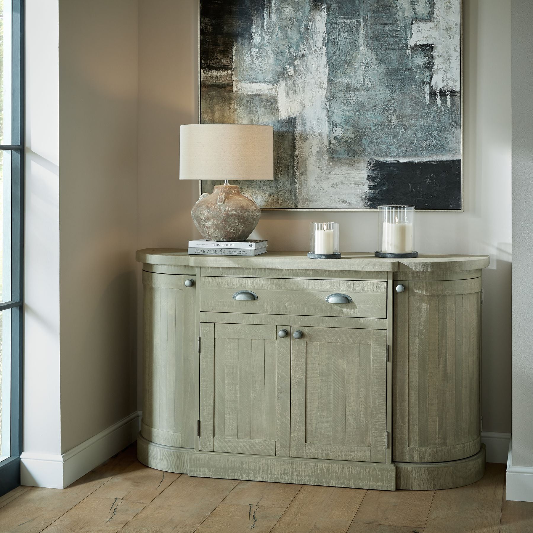 Wooden sideboard with lamp and candles in a room with abstract art on the wall.