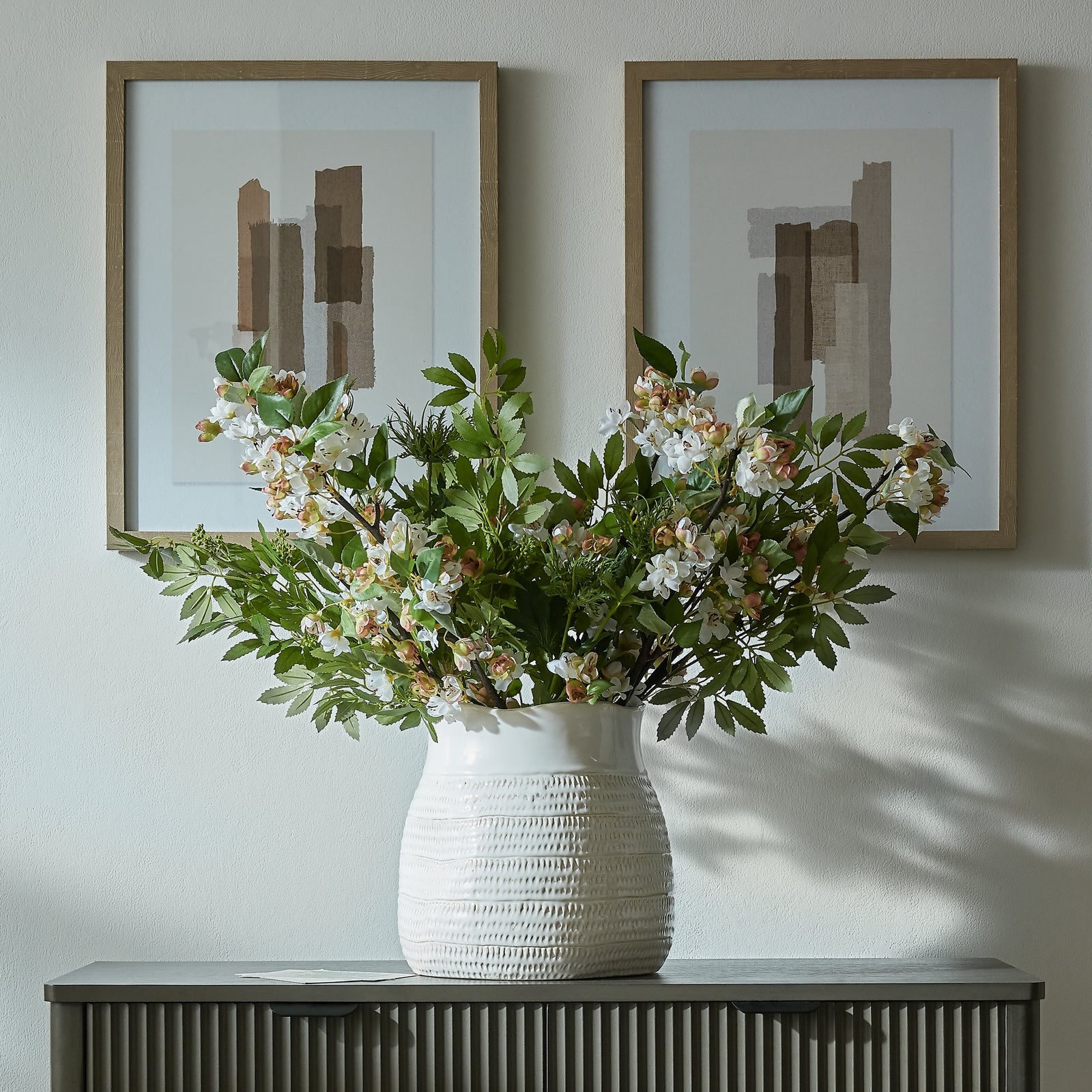 White vase with flowers on a console table against a wall with two framed abstract artworks.