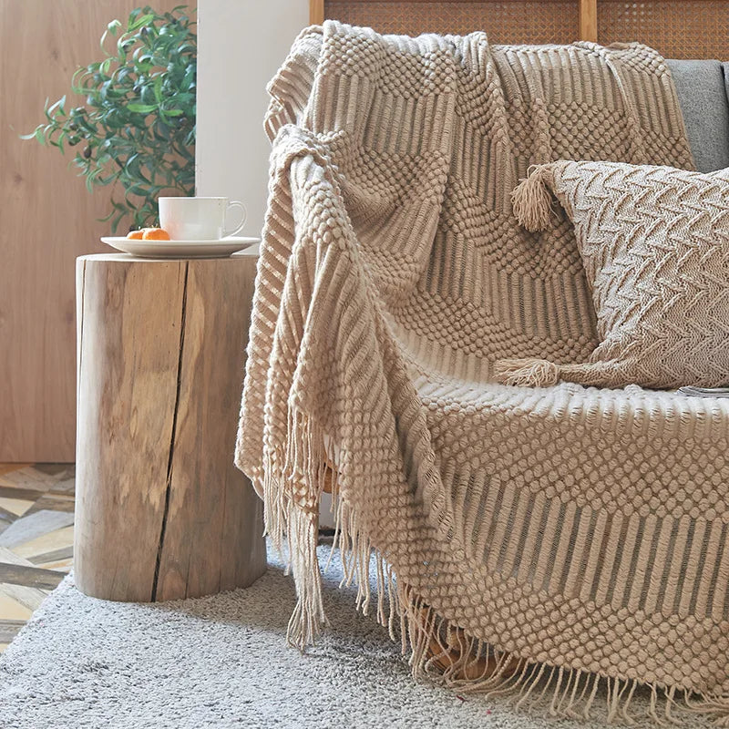 Beige knitted blanket draped over a sofa with a wooden side table and plant in the background.