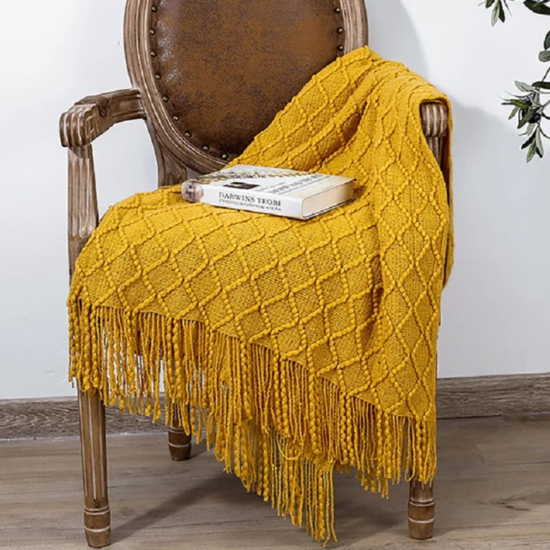 Yellow knitted blanket with fringes draped over a wooden chair with a book on it.