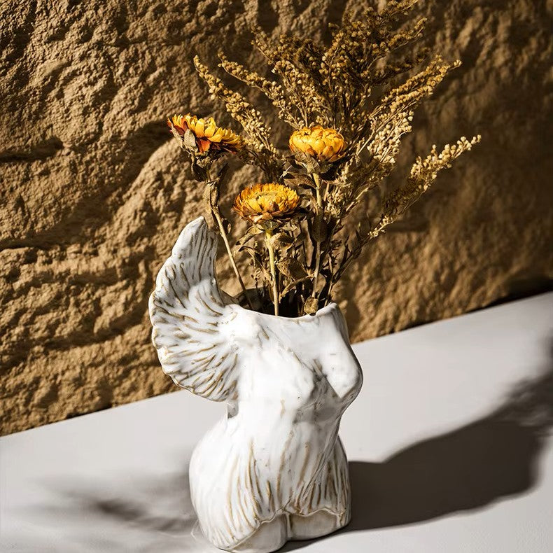 Vase shaped like a woman's torso with dried flowers against a stone wall.