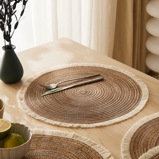 Round woven placemats with fringe on a wooden table, set with cutlery.