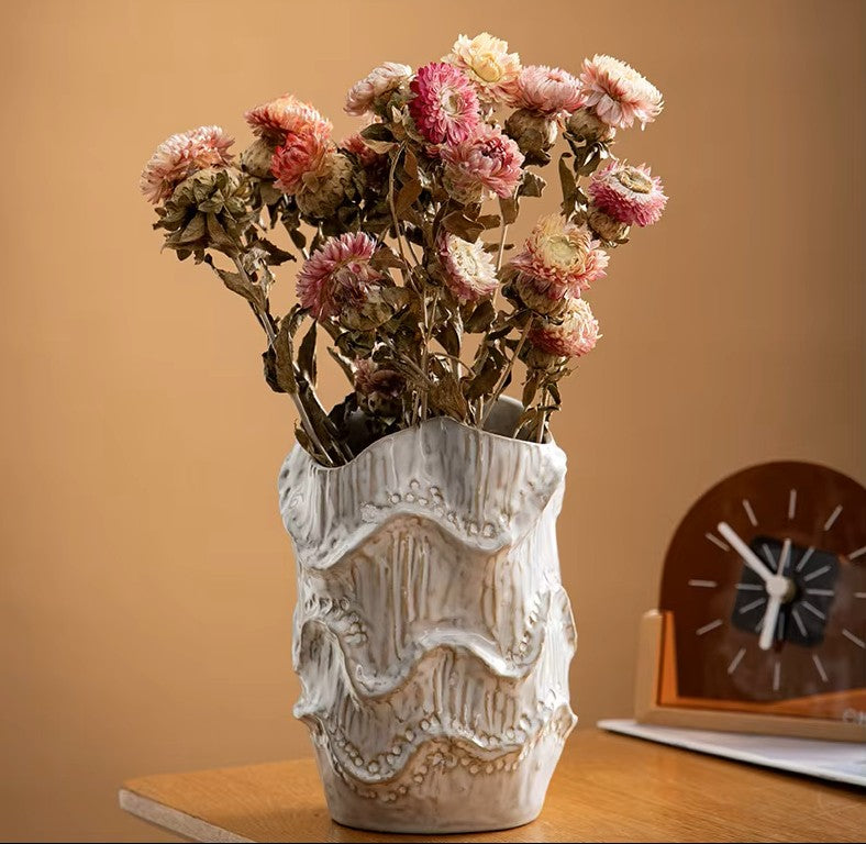 Decorative vase with dried flowers on a wooden surface against a beige wall.