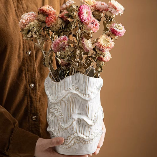 Person holding a decorative white vase with dried pink flowers against a brown background